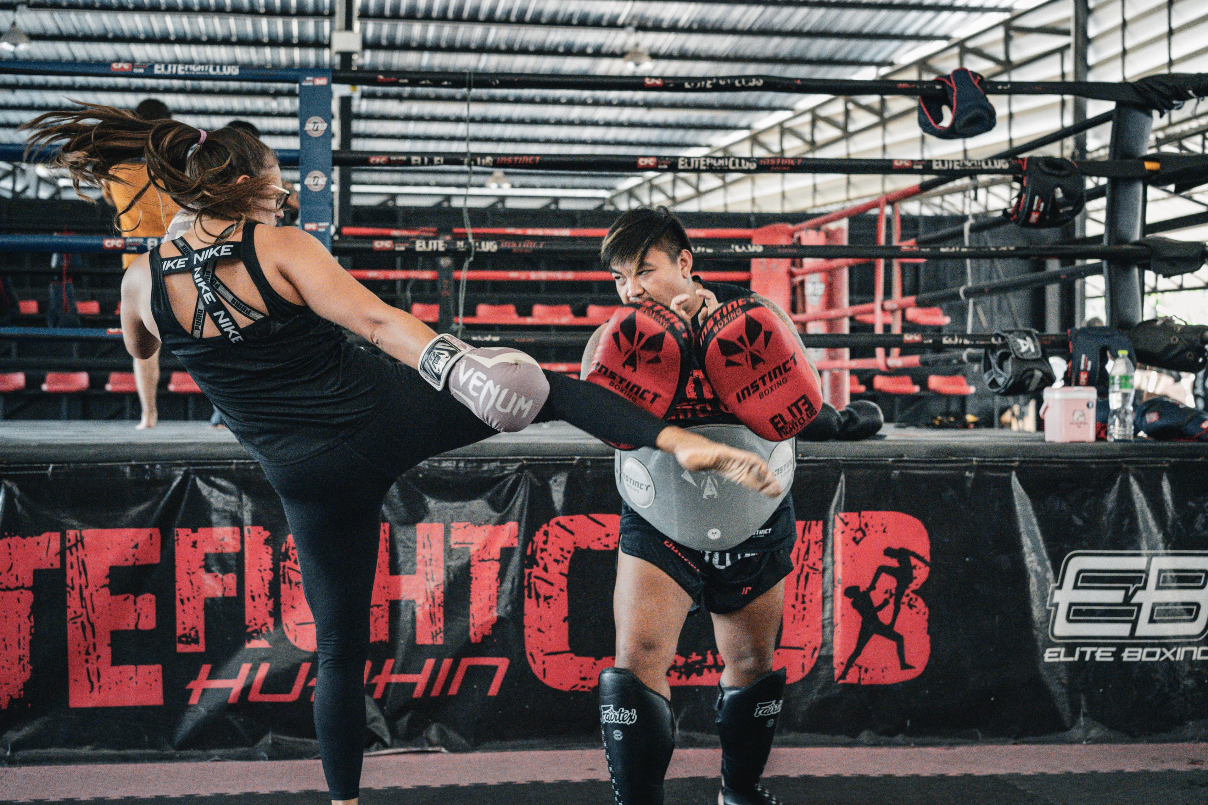 Au camp, une participante frappe d’un coup de pied les paos tenus par un entraîneur, devant la bannière Elite Fight Club.